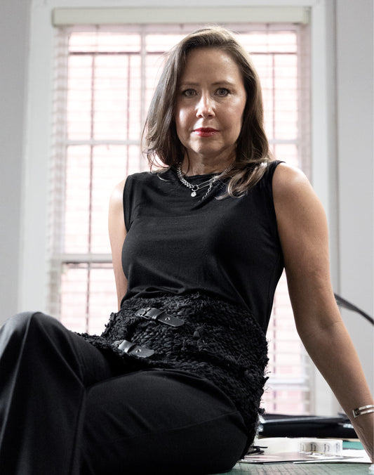 A woman with shoulder-length brown hair sits indoors wearing a sleeveless black top and black pants, with a textured black belt and layered silver necklaces. She is posed in front of a window with blinds.